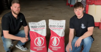 Cole the Cornstar and Brandon Hulme kneeling on the dirt floor of a barn with two bags of Champion Seed seed corn in between them