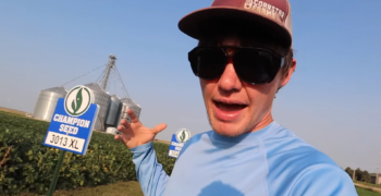 Cole the Cornstar standing in front of a soybean field with Champion Seed field signs in the background