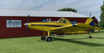 Yellow plane with blue stripe getting ready to fly from a hangar on a farm that includes the Champion Seed logo on the barn.